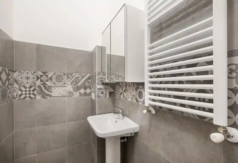 Compact modern bathroom with a pedestal sink, mirrored cabinet and a vertical heated towel rail; patterned gray tiles add decorative detail.