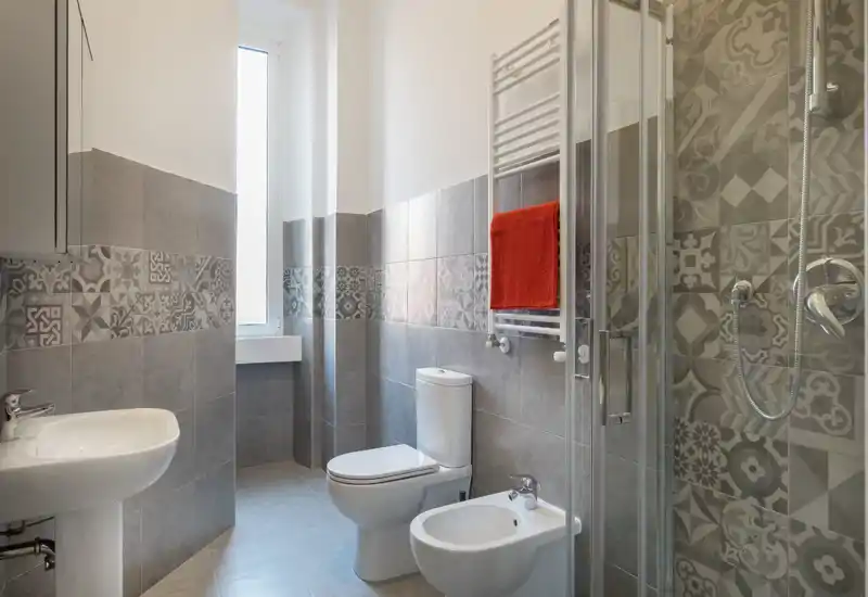 Bright, modern bathroom with patterned grey tiles, a shower with glass door, toilet, bidet and sink; a tall window provides natural light and a red towel adds a pop of color.