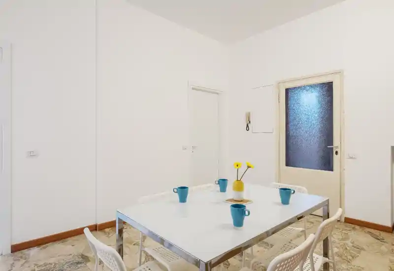 Minimalist dining area with a white rectangular table, six perforated chairs and a small central vase with yellow flowers; bright neutral walls and decorative blue cups add contrast.