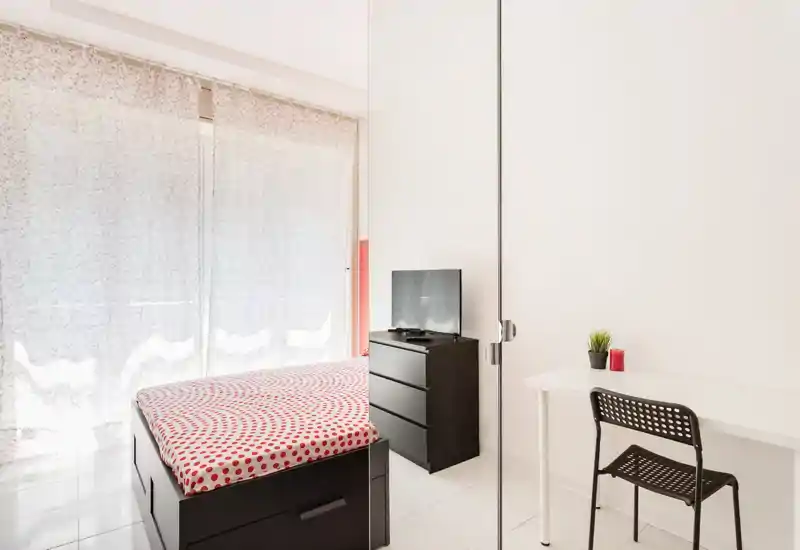 Dormitorio moderno y luminoso con cama doble con funda de lunares rojos, cómoda negra con TV y un escritorio blanco sencillo. Gran ventana con cortinas translúcidas que aporta mucha luz natural, creando un ambiente limpio y minimalista ideal para alquileres temporales.