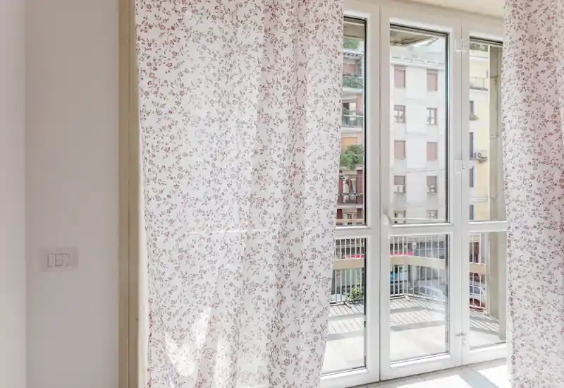 Acceso a balcón/terraza luminoso a través de grandes puertas de cristal con cortinas transparentes estampadas; se ven edificios y la barandilla exterior.