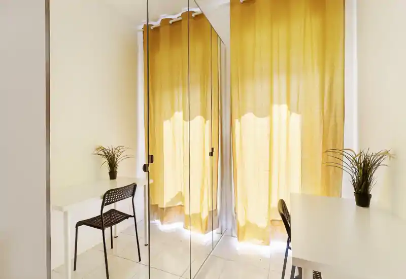 Espacio diurno luminoso y minimalista con dos escritorios, sillas y un armario con espejo que refleja cortinas amarillas; pequeñas plantas aportan un toque verde.