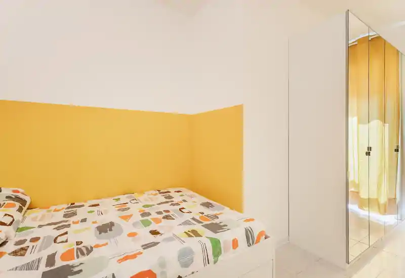 Dormitorio compacto y soleado con cama doble de ropa de cama con estampado y pared amarilla; espacio limpio con armario con espejo y agradable luz natural.