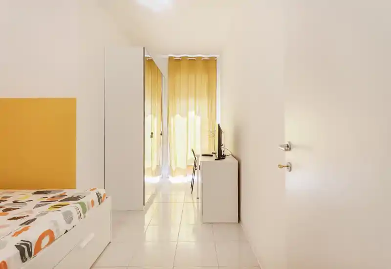 Dormitorio luminoso y minimalista con muebles blancos, ropa de cama estampada y cortinas amarillas que dejan pasar luz natural suave.
