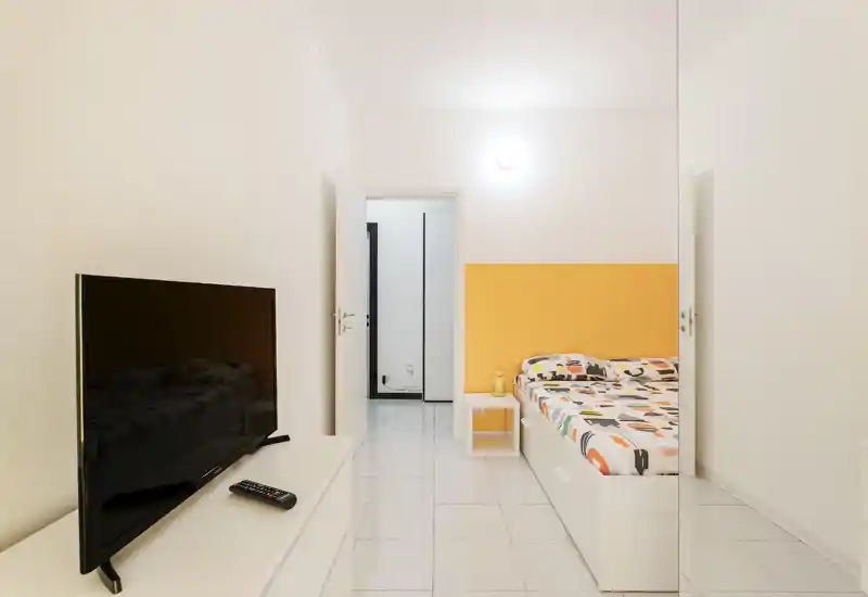 Dormitorio luminoso y minimalista con cama individual/mediana junto a una pared amarilla, mesita ordenada y televisor enfrente — sencillo y acogedor.