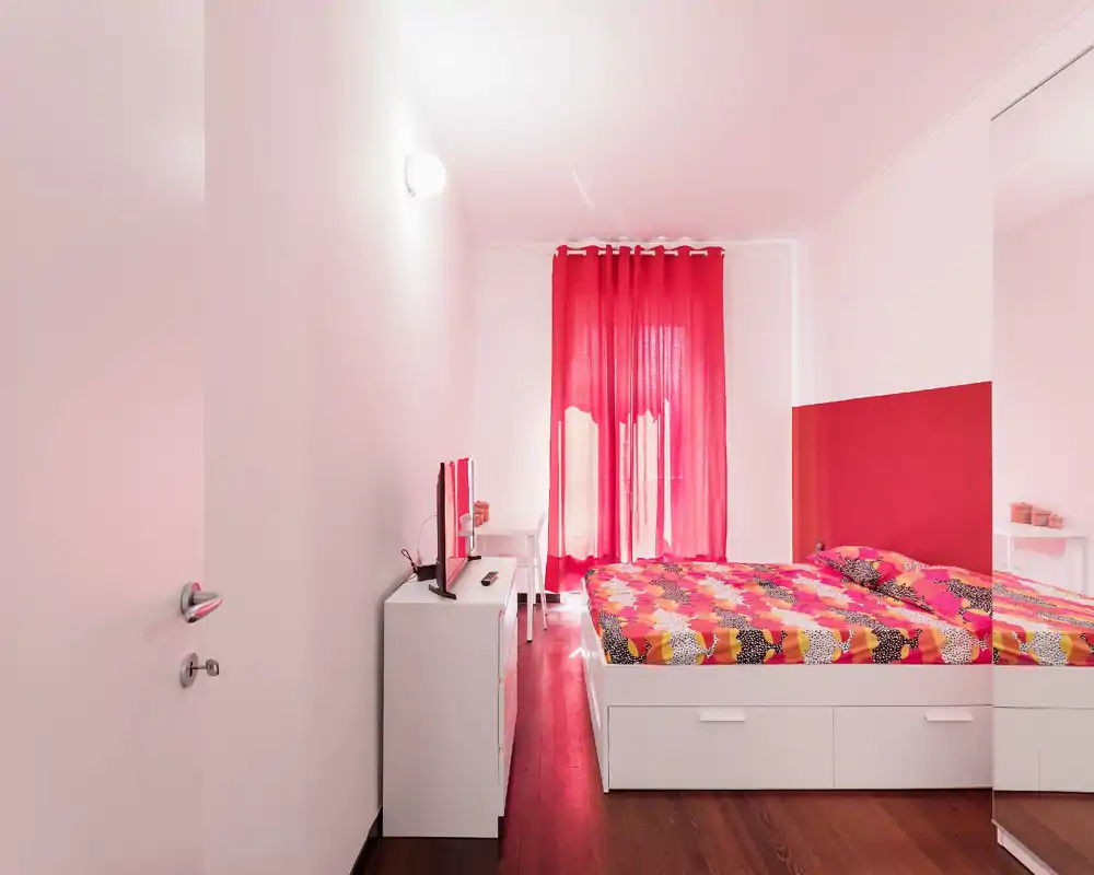 Bright, modern bedroom with a colorful double bed, white storage drawers and a wall-mounted TV. The room features vibrant red curtains and a matching accent wall, creating a lively yet cozy atmosphere.