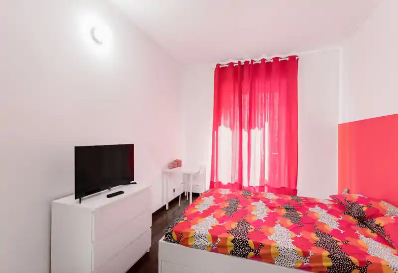 Bright, compact bedroom with a patterned double bed, red curtains and a white dresser with TV — cozy, well-lit and stylish for short stays.