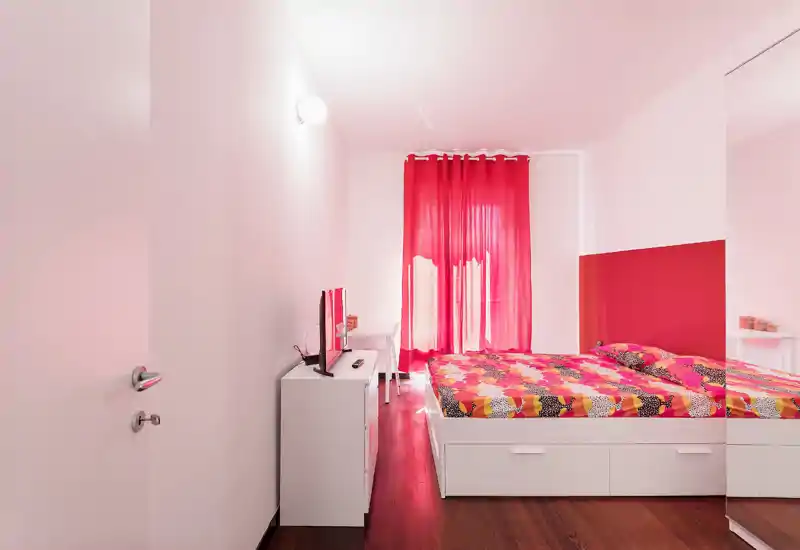 Bright, modern bedroom with a colorful double bed, white storage drawers and a wall-mounted TV. The room features vibrant red curtains and a matching accent wall, creating a lively yet cozy atmosphere.