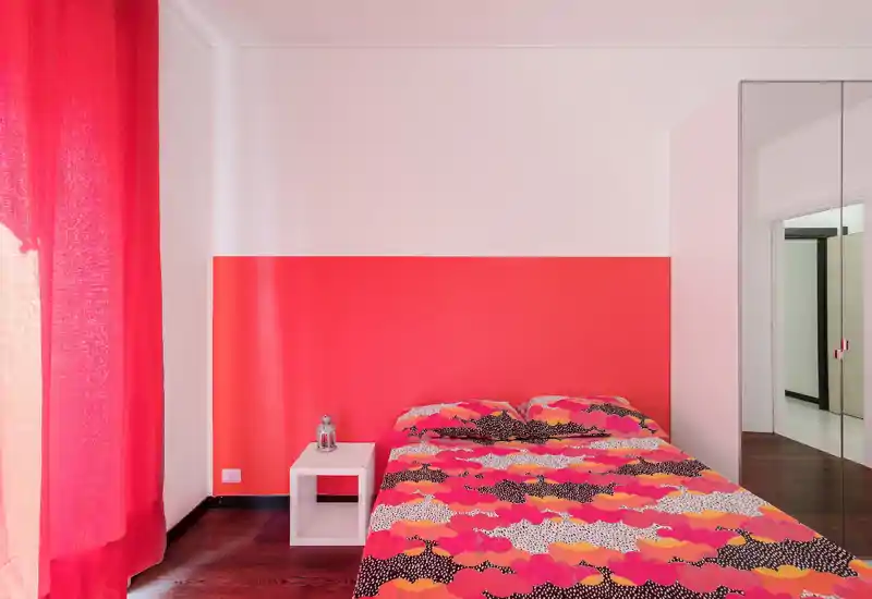 Bright, stylish bedroom with bold coral accent wall and patterned bedding; features a simple bedside table and natural light from large curtains.