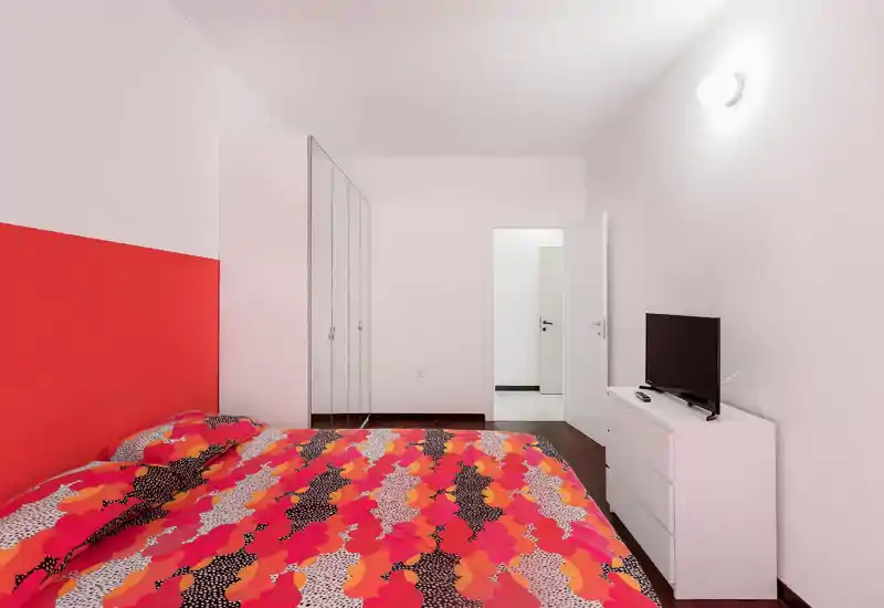 Bright, modern bedroom with a large bed featuring colorful bedding, a white dresser with TV, and built-in mirrored wardrobe — clean minimalist styling ideal for rentals.