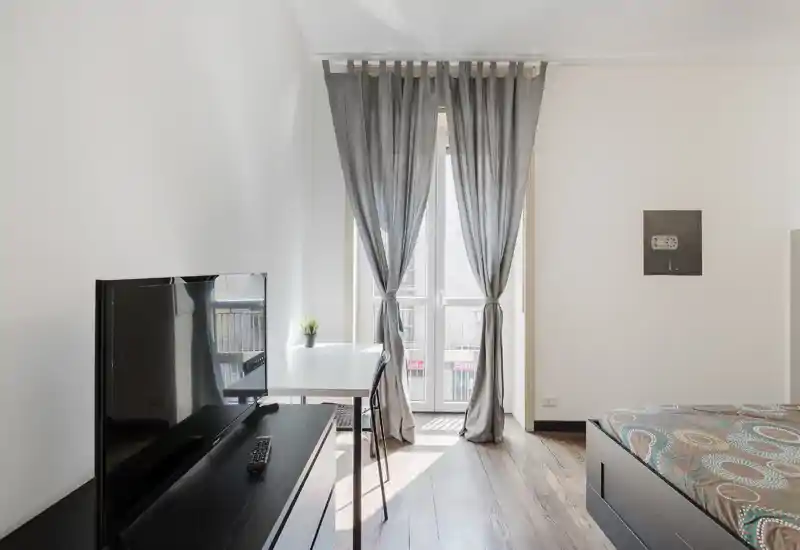 Dormitorio luminoso y minimalista con la cama visible a la derecha, televisor sobre un mueble bajo y un pequeño escritorio junto a una ventana alta con cortinas que dejan entrar la luz natural.