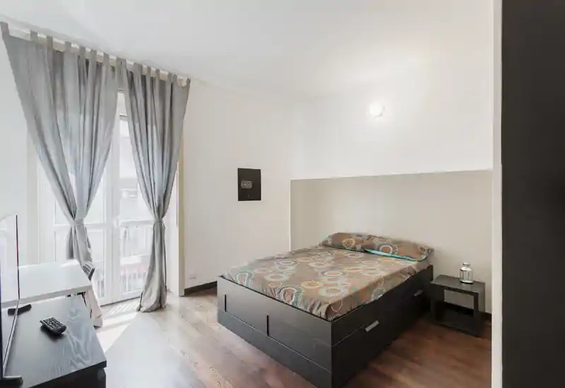 Dormitorio doble moderno y luminoso con cama tipo plataforma negra, ropa de cama con estampado y luz natural desde el balcón. Líneas limpias y suelo de madera que lo hacen acogedor y fácil de amueblar.