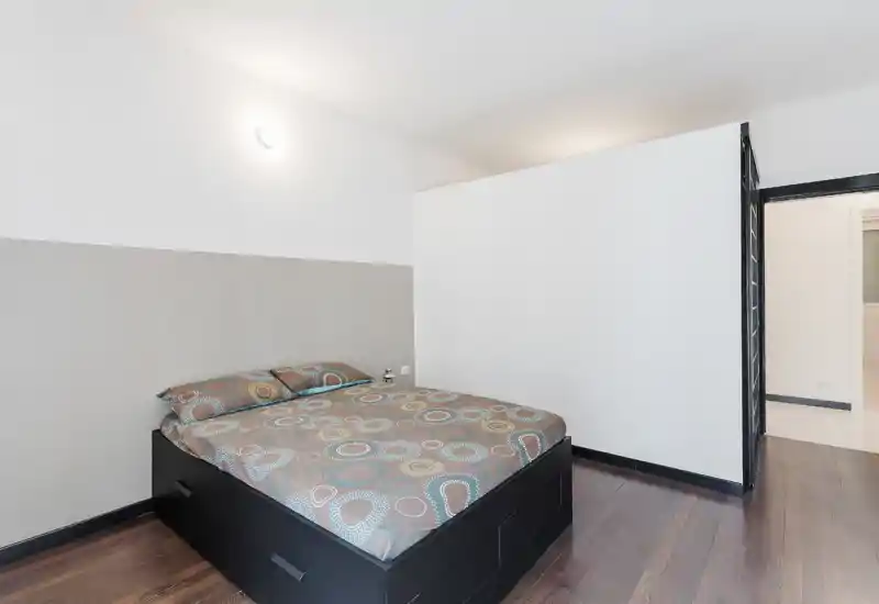 Dormitorio moderno y luminoso con cama doble en estructura baja negra, ropa de cama estampada y suelos de madera cálida. Estilo minimalista y limpio con luz de pared suave y tonos neutros.