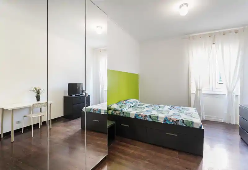 Camera da letto luminosa e moderna con letto matrimoniale, armadio con ante a specchio, pavimenti in legno e scrivania vicino alla finestra. Arredamento pulito e minimalista con luce naturale, ideale per gli annunci.