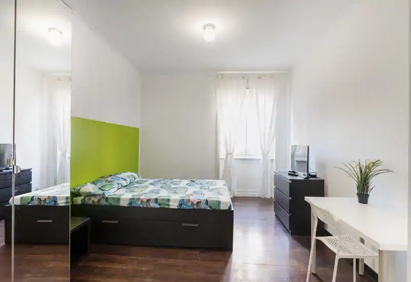 Camera da letto luminosa e moderna con letto matrimoniale su contenitore scuro, pareti bianche con una striscia verde, tende leggere e una piccola scrivania con TV—pulita, minimale e ben illuminata.