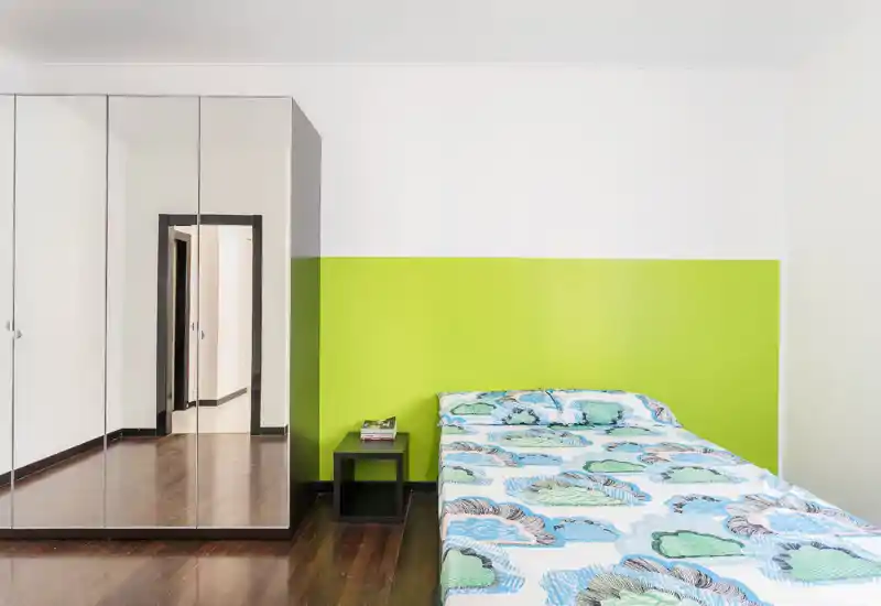 Camera da letto luminosa e minimal con letto matrimoniale contro una parete lime, armadio con ante a specchio, pavimento in legno e un piccolo comodino.