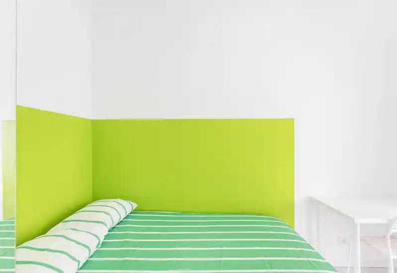Dormitorio minimalista y luminoso con cabecero verde y colcha de rayas verdes; se aprecia un escritorio sencillo a un lado.