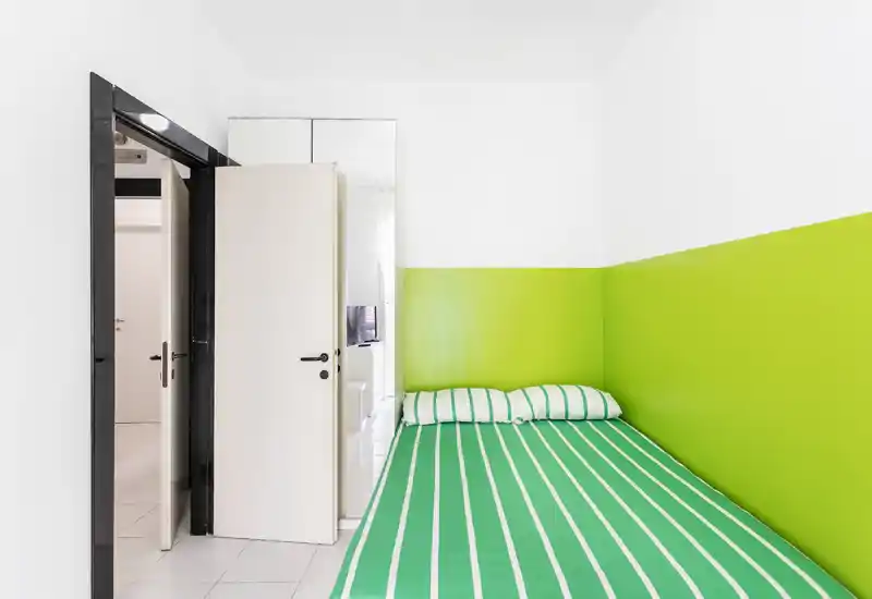 Dormitorio compacto y luminoso con pared verde y ropa de cama a rayas a juego. La cama ocupa el primer plano; ambiente fresco y minimalista, ideal para una persona o estancias cortas.