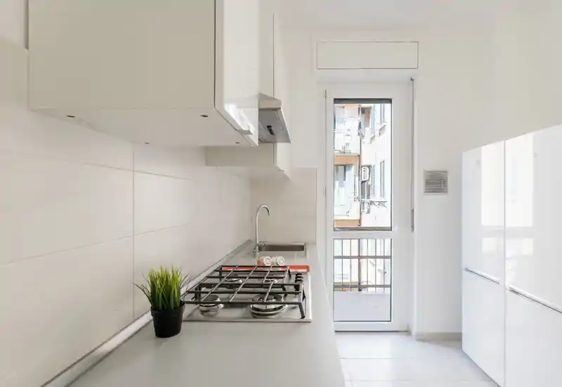 Cucina moderna e luminosa a corridoio con piano cottura a gas, cappa integrata, mobili bianchi e porta sul balconcino. Superfici minimal e una piccola pianta decorativa rendono lo spazio ordinato e funzionale.