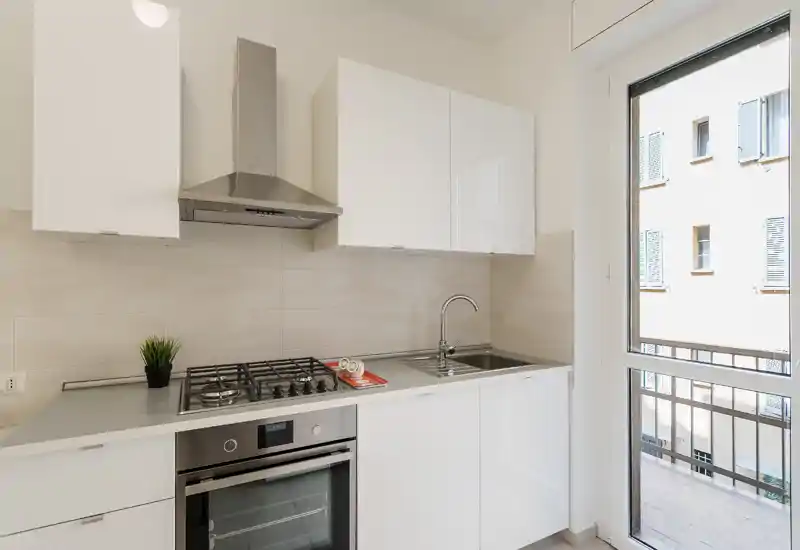 Cucina moderna e compatta con mobili bianchi, forno in acciaio inox e piano cottura a gas, lavello ben illuminato dalla luce naturale e porta che si apre su un balcone.