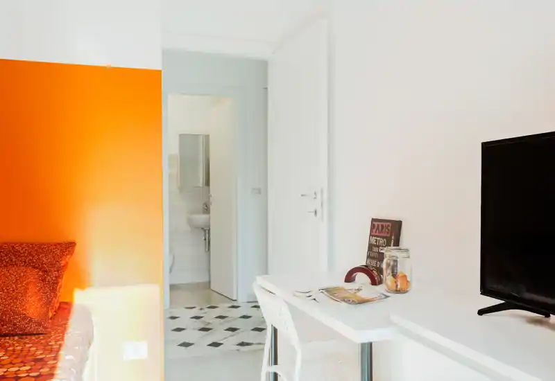 Bright, modern bedroom corner with an orange accent wall and a bed with patterned textiles; a white desk and TV sit opposite, and a doorway leads to the bathroom.