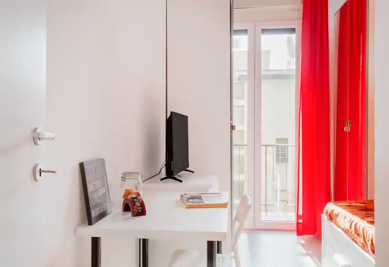 Compact, bright bedroom corner with a partially visible bed, desk with a TV and a small balcony letting in natural light through vivid red curtains.