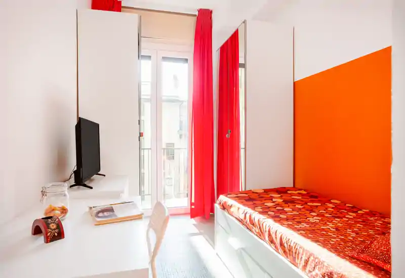Compact, bright bedroom with a single bed dressed in a warm patterned cover, bold orange accent wall and red curtains that frame a balcony door. A white desk with a chair and TV sits opposite the bed, offering a practical study corner.