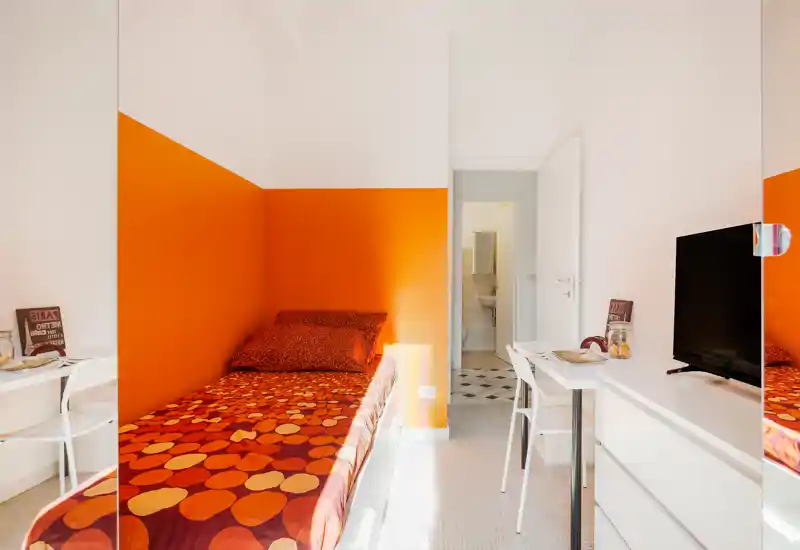 Compact, brightly lit single bedroom with warm orange accent wall and patterned bedding. The space includes a desk, chair and TV, ideal for a student or short-term stay.