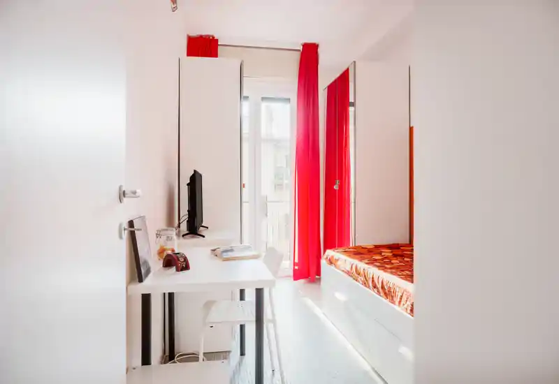 Compact bright bedroom with a single bed, built-in storage drawers and a small desk by a sunny balcony. The vivid red curtains and warm orange bedspread create a lively, modern look ideal for rental photos.