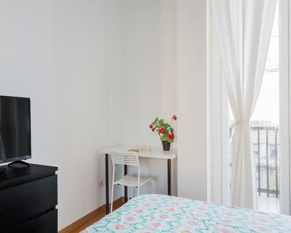 Bright, minimal bedroom with patterned bedspread, a small desk and chair, TV dresser and a balcony window letting in natural light.