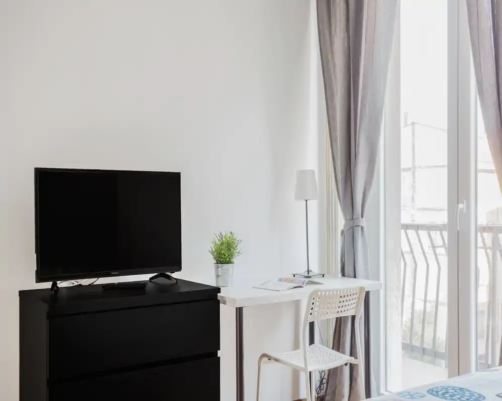 Angolo camera da letto luminoso e minimalista con scrivania, TV su comò e un piccolo lembo del letto vicino alla finestra; toni neutri e luce naturale per un ambiente arioso.