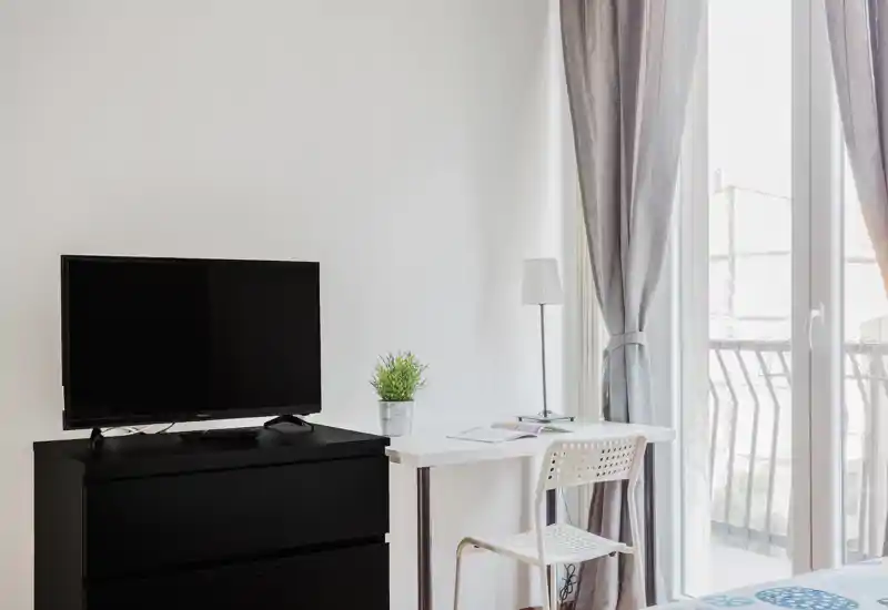 Angolo camera da letto luminoso e minimalista con scrivania, TV su comò e un piccolo lembo del letto vicino alla finestra; toni neutri e luce naturale per un ambiente arioso.