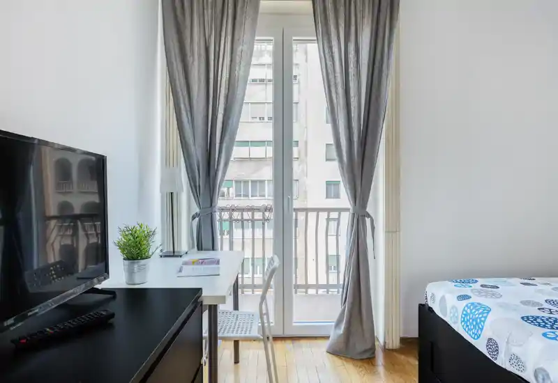 Angolo camera da letto luminoso e moderno con letto singolo, scrivania e porte sul balcone; arredi neutri e luce naturale rendono lo spazio arioso e funzionale, ideale per una persona o affitti brevi.