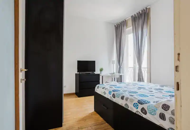Camera da letto moderna e luminosa con letto matrimoniale con copriletto fantasia, comò con TV e una piccola scrivania vicino alle ampie finestre che lasciano entrare luce naturale.