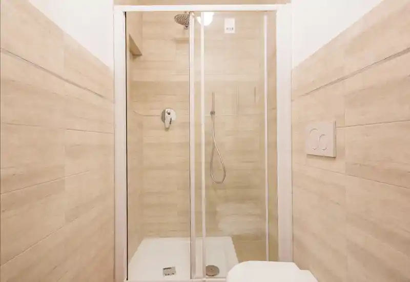 Bagno moderno compatto con box doccia in vetro, pareti rivestite in piastrelle beige e pannello di scarico a parete. Design pulito e minimal, ideale per affitti brevi.