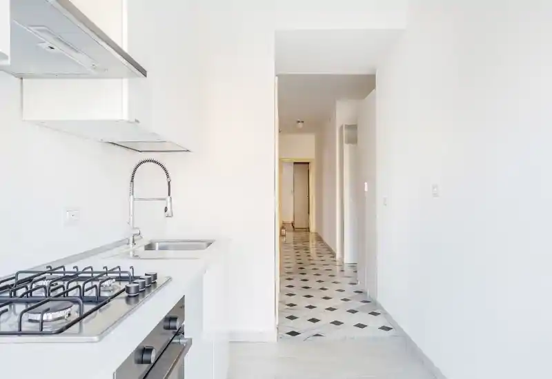 Bright modern kitchen with a stainless-steel sink, gas cooktop and minimalist white cabinetry; clean, airy layout with a tiled hallway visible beyond.