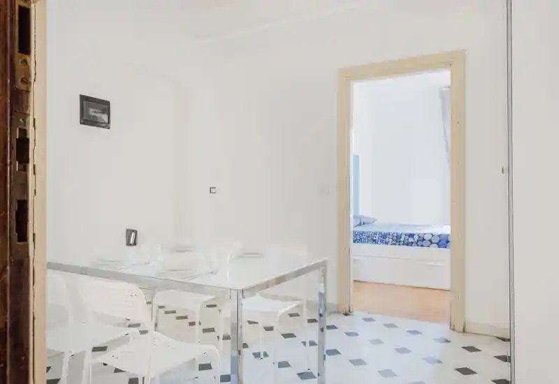 Minimalist dining area with a glass/metal table and white chairs on patterned tile, set for a meal; a doorway reveals a bedroom beyond.