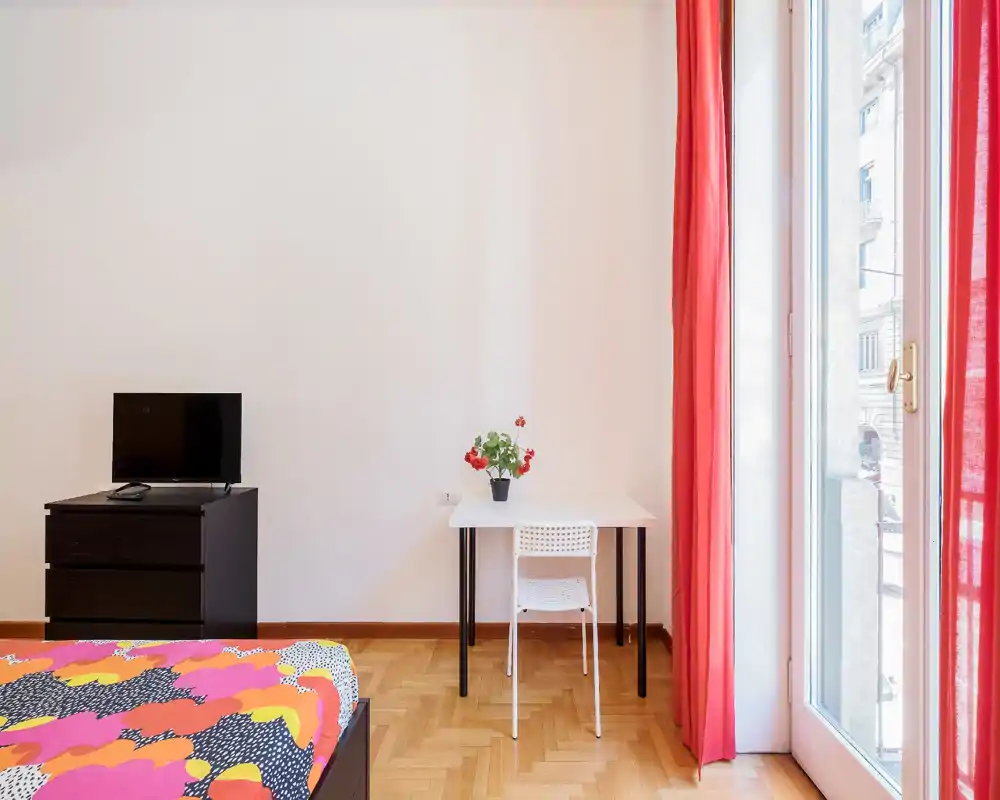 Camera da letto luminosa e minimale con una coperta colorata, una piccola scrivania con sedia e vaso, TV su un comò basso e una grande finestra con tende rosse che lasciano entrare molta luce naturale.