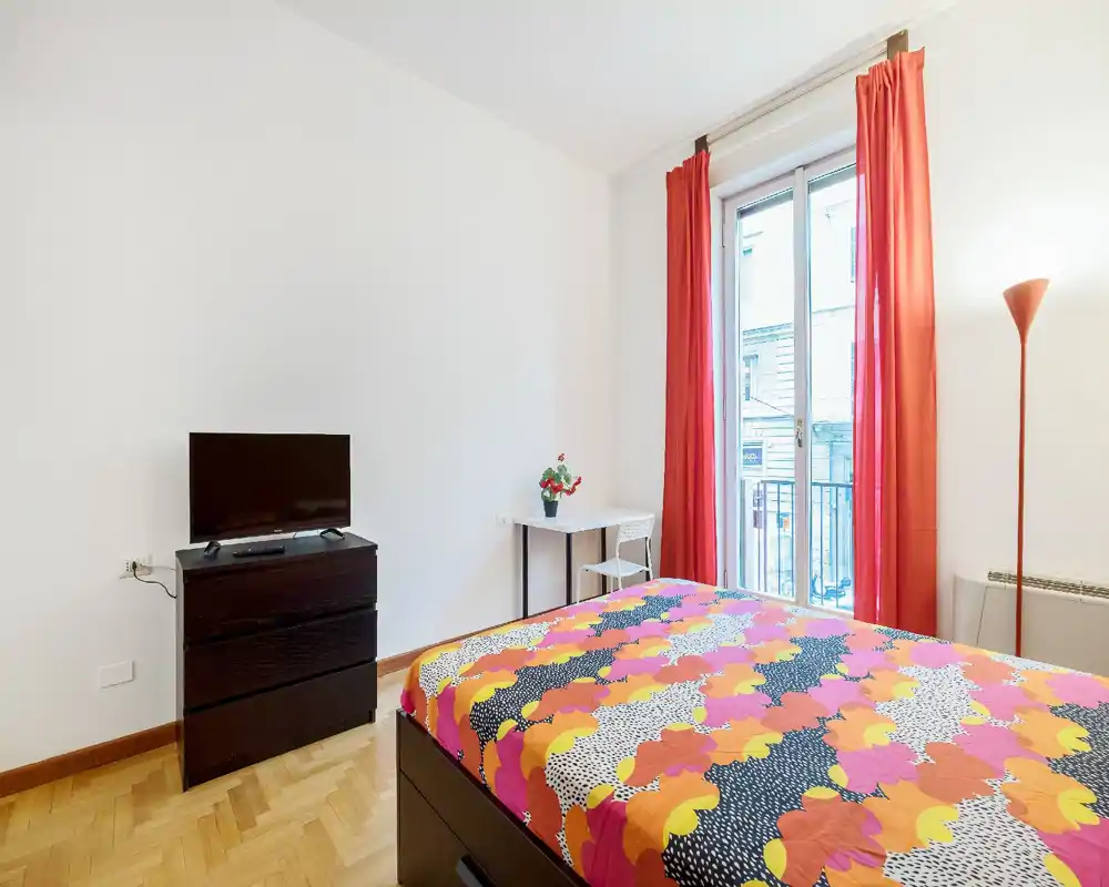 Camera da letto luminosa e colorata con letto matrimoniale in primo piano, ampia finestra con tende rosse, piccolo scrittoio e TV — ben illuminata e accogliente.