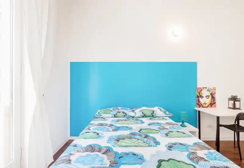 Camera da letto luminosa e moderna con letto matrimoniale e biancheria colorata davanti a una testata turchese; luce naturale da una grande finestra e una scrivania semplice rendono l'ambiente arioso.