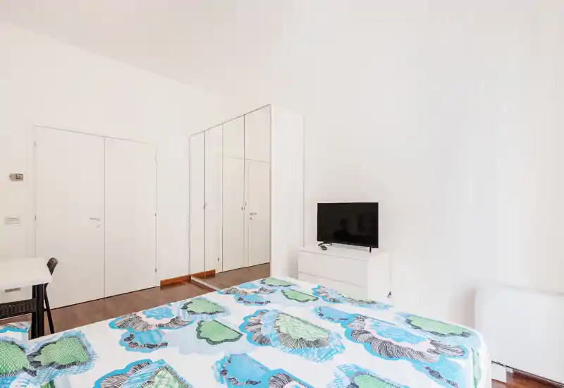 Camera da letto luminosa e minimalista con letto matrimoniale con copriletto colorato blu-verde, armadio con ante a specchio, cassettiera con TV e una semplice scrivania — perfetta per un affitto compatto.