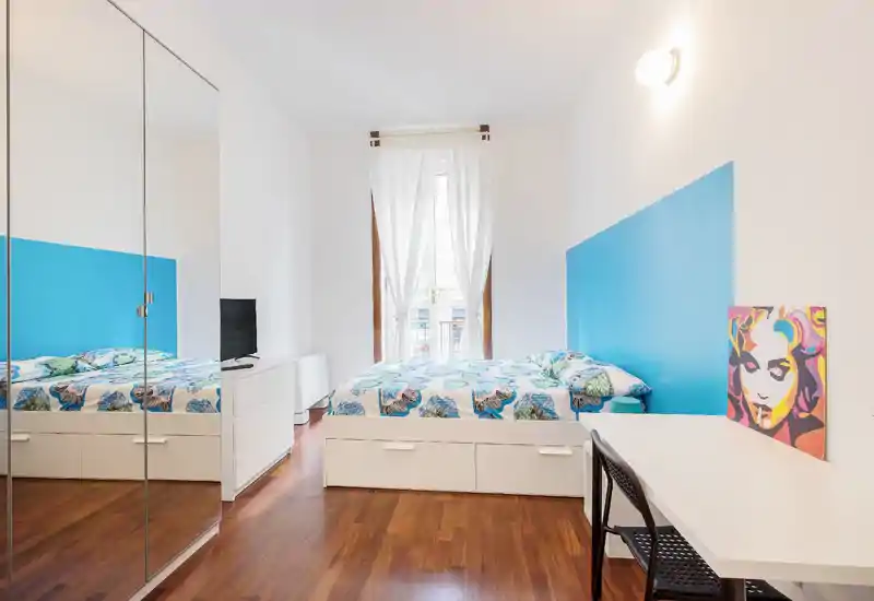 Camera da letto luminosa e moderna con letto matrimoniale, biancheria colorata e grande armadio con ante specchiate che dà profondità. Mobili bianchi e pavimento in legno creano un aspetto pulito e arioso con molta luce naturale.