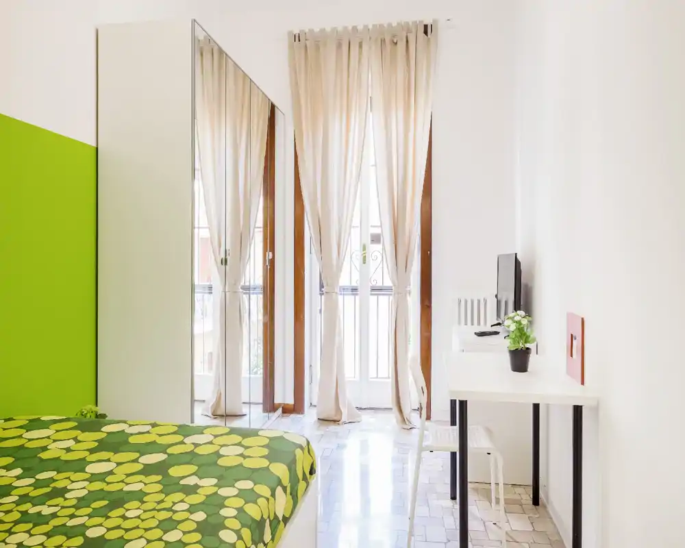 Camera da letto luminosa e moderna con copriletto verde a fantasia, armadio con specchio a tutta altezza e balcone illuminato da tende leggere.