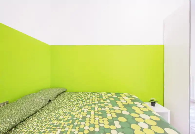 Dormitorio luminoso y moderno con una cama grande cubierta con ropa de cama verde y amarilla con estampado, pared bicolor verde lima y blanca, decoración mínima con una mesita y una planta.