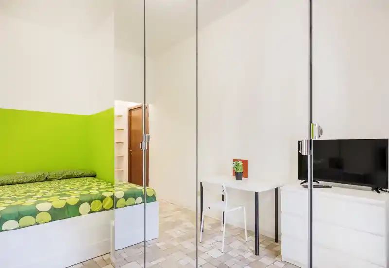 Dormitorio moderno y luminoso con pared de acento verde y ropa de cama estampada, pequeño escritorio y TV: espacio compacto, bien iluminado y ordenado.