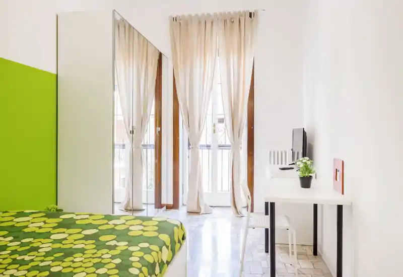 Dormitorio moderno y luminoso con colcha verde estampada, armario con espejo de cuerpo entero y balcón con cortinas translúcidas.