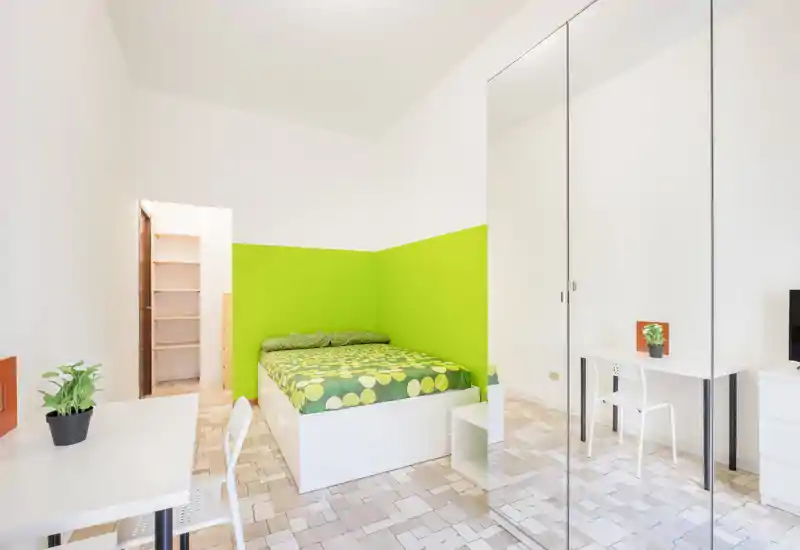 Dormitorio moderno y luminoso con cama elevada frente a una pared verde brillante, armario con espejo y escritorios minimalistas — ideal para estudiante o persona sola.