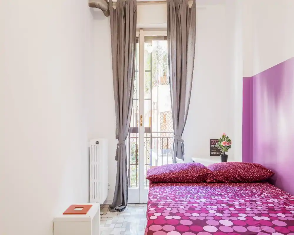 Camera da letto compatta e luminosa con un letto matrimoniale coperto da biancheria fucsia, una parete d'accento viola e luce naturale da una finestra alta con tende grigie. Comodino semplice e piccola pianta rendono l'ambiente accogliente e adatto ad affitti brevi.
