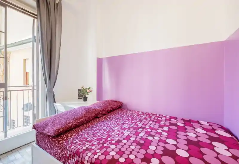 Camera da letto compatta e luminosa con letto completamente visibile e copriletto rosa a pois, luce naturale dalla porta del balcone e una piccola scrivania con pianta — ideale per una singola ospitalità vivace.