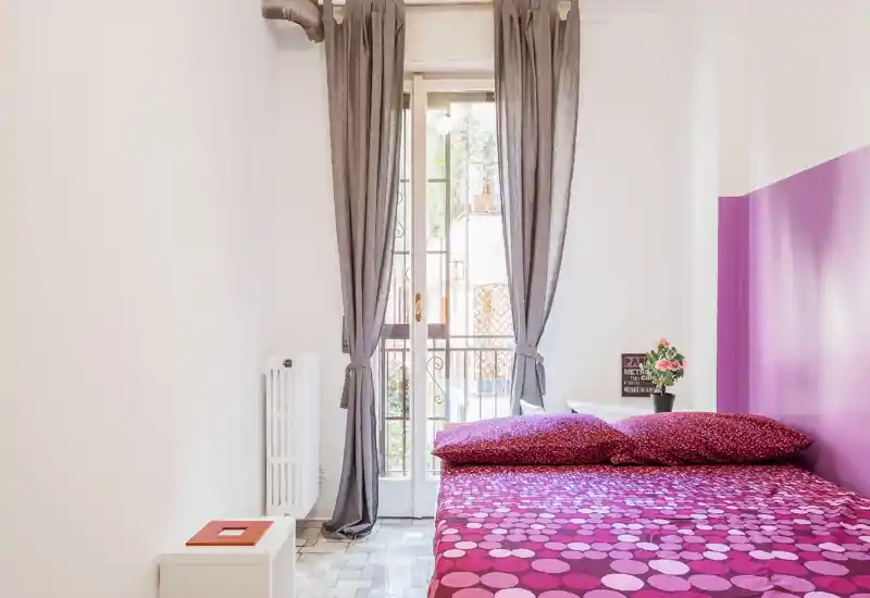 Camera da letto compatta e luminosa con un letto matrimoniale coperto da biancheria fucsia, una parete d'accento viola e luce naturale da una finestra alta con tende grigie. Comodino semplice e piccola pianta rendono l'ambiente accogliente e adatto ad affitti brevi.
